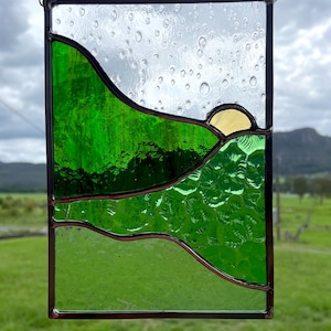 May include: Stained glass panel depicting a landscape scene with green hills, a yellow sun, and a clear sky. The glass is framed in a dark metal and suspended by a chain. The design evokes a sense of nature and tranquility.
