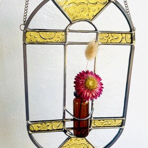 May include: A decorative stained glass window with a metal frame and chain for hanging. The window features clear glass panes with yellow patterned accents and a small brown bottle holding a pink flower and dried grass.