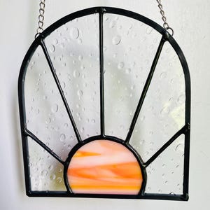 May include: A stained glass sun catcher with a black metal frame and a pink and orange sunset design. The glass is textured with small bubbles.