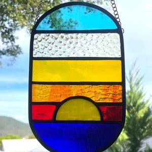 May include: Stained glass panel depicting a sunset scene. The panel features a blue sky, textured white clouds, and bands of yellow, orange, and red, with a yellow sun and a blue sea. The panel is framed in black and hangs from a chain.