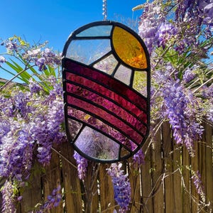 May include: An oval stained glass suncatcher featuring a black metal frame. The design includes a yellow sun in the upper right corner, with horizontal bands of textured magenta and purple glass below, and a clear glass section at the bottom.