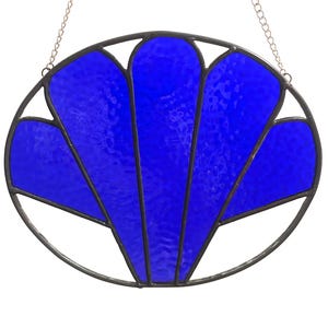 May include: A stained glass wall hanging with a vibrant blue fan-shaped design. The glass is set within a dark metal frame and suspended by a silver chain. The design is reminiscent of Art Deco style.