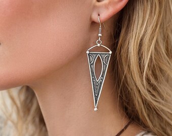May include: Silver-toned, triangular drop earrings with an open oval design in the center. The earrings have intricate etched patterns and a small ball at the bottom. The earrings are hanging from a silver hook.