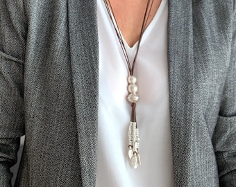 May include: A long, brown leather necklace with silver beads and a tassel pendant. The necklace is worn over a white top and a grey herringbone blazer.