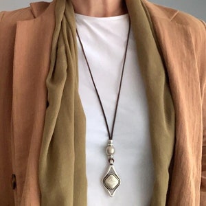 Peut inclure: Un collier en cordon de cuir marron avec un pendentif argenté orné d'une pierre blanche et d'une perle ronde. Le collier est porté par une personne vêtue d'une chemise blanche et d'une veste marron.