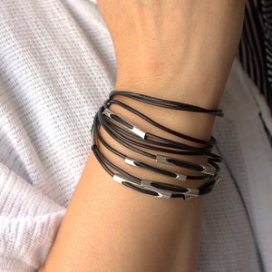 May include: A multi-strand bracelet featuring thin black leather cords wrapped multiple times around the wrist. The design incorporates several small, smooth, silver-toned metal cylindrical beads spaced evenly along the leather strands.