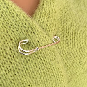Könnte beinhalten: Eine silber-goldene Sicherheitsnadel, die einen hellgrünen Pullover befestigt. Die Nadel hat einen gebogenen silbernen Kopf und einen geraden goldenen Stift. Der Pullover ist aus weichem Strickmaterial.