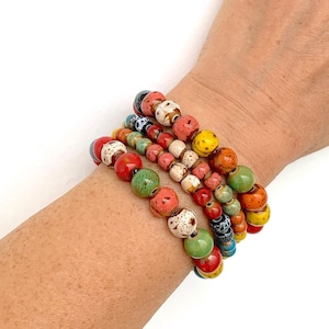 Könnte beinhalten: Ein Set aus fünf bunten Keramikperlenarmbändern. Die Armbänder sind auf dem Handgelenk einer Person gestapelt und zeigen eine Vielzahl von Farben, darunter Rot, Orange, Gelb, Grün, Blau und Weiß. Die Perlen sind mit verschiedenen Farben gesprenkelt und haben einen einzigartigen, handgefertigten Look.