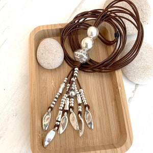 K&ouml;nnte beinhalten: Eine lange braune Lederkette mit silberfarbenen Perlen und blattf&ouml;rmigen Anh&auml;ngern. Die Kette wird auf einem Holztablett mit zwei glatten Steinen pr&auml;sentiert. Die Kette hat einen Bohemian-Stil.