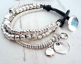 May include: A multi-strand bracelet featuring rows of metallic silver-toned beads strung on black leather cords. The bracelet includes a large circular silver-toned button clasp and two dangling circular silver-toned charms at the end.