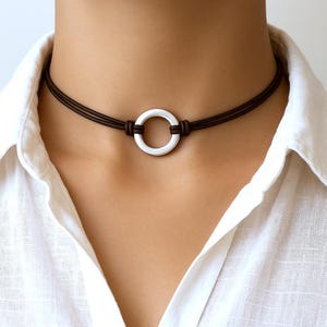 Puede incluir: Un collar tipo gargantilla de cordón marrón oscuro con un anillo circular plateado en el centro. El cordón consta de tres hilos finos anudados firmemente a ambos lados del anillo de metal, sobre una camisa blanca con cuello.