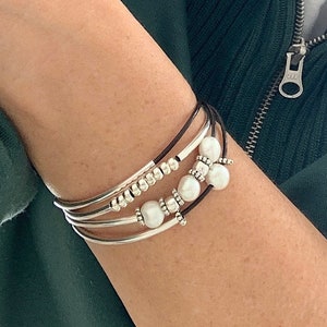 Könnte beinhalten: Ein Set aus vier Armbändern mit silbernen Perlen und weißen Perlen. Die Armbänder sind übereinander am Handgelenk einer Person gelegt.