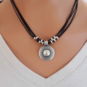 May include: A multi-strand black cord necklace featuring a circular silver-toned pendant with a central spiral design and radiating etched lines. The pendant is accented by small silver-toned cylindrical beads on each side of the cord strands.