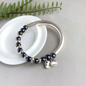 Pode incluir: Um bracelete de prata com uma fila de contas pretas iridescentes e dois pingentes de cora&ccedil;&atilde;o prateados.