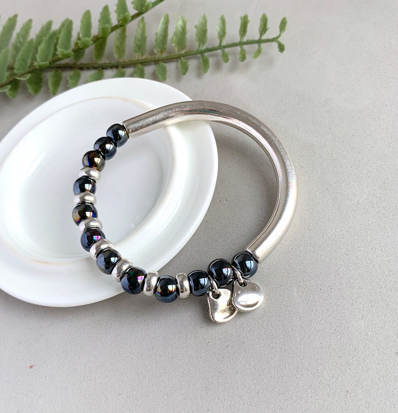 Pode incluir: Um bracelete de prata com uma fila de contas pretas iridescentes e dois pingentes de cora&ccedil;&atilde;o prateados.