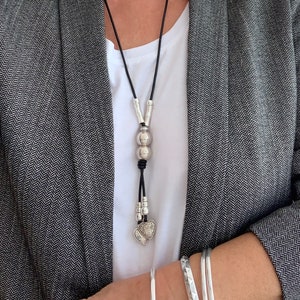 May include: A silver necklace with a heart pendant and two round beads. The necklace is made of black leather cord and has a silver clasp.