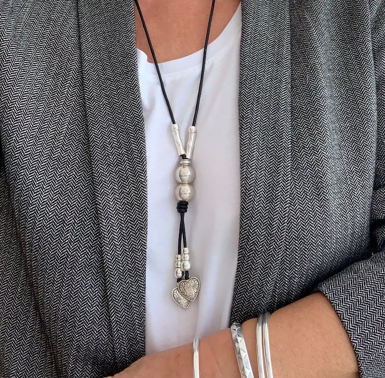 May include: A silver necklace with a heart pendant and two round beads. The necklace is made of black leather cord and has a silver clasp.