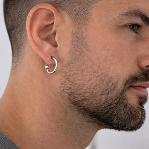 Pode incluir: Um brinco de argola prateado com um pequeno detalhe de bola. O brinco é usado na orelha. A pessoa na imagem tem um corte de cabelo curto e barba.