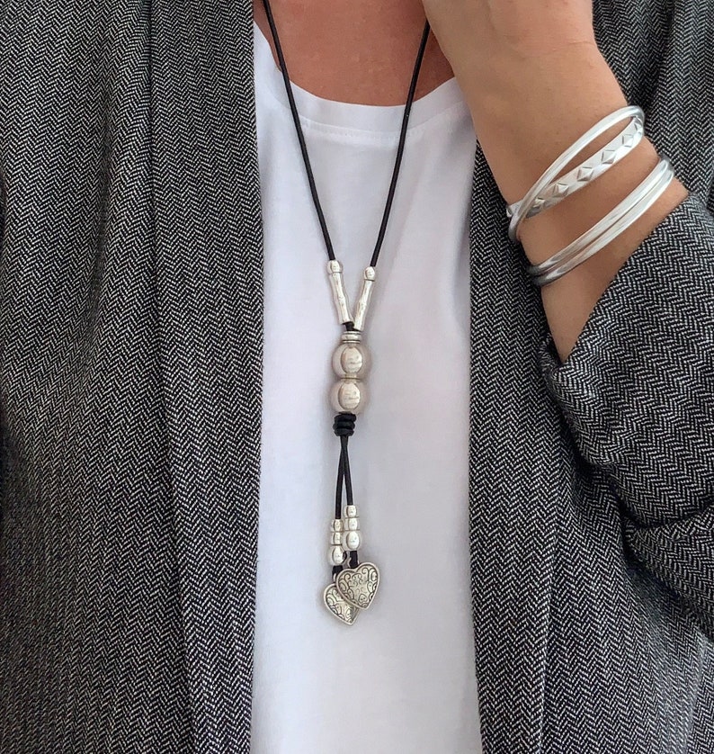 May include: A silver necklace with a leather cord and two heart charms. The necklace also features a silver bead with a textured surface.