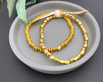 Può includere: Due braccialetti dorati con perline su una superficie grigia. I braccialetti sono fatti di piccole perline di forma irregolare.