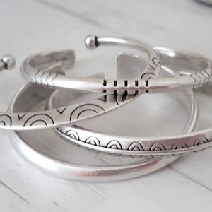 Op de afbeelding: Een set van drie zilverkleurige metalen armbanden. De bovenste armband heeft een open ontwerp met bolvormige uiteinden en gegraveerde horizontale lijnen en golfpatronen. De middelste armband toont een halve cirkelpatroon, de onderste is glad.