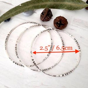May include: Three silver bangles with a textured design, each measuring 2.5 inches in diameter. The bangles are displayed on a white surface, with natural elements in the background.