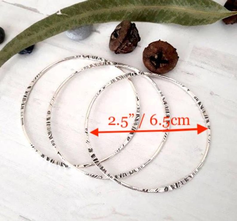 May include: Three silver bangles with a textured design, each measuring 2.5 inches in diameter. The bangles are displayed on a white surface, with natural elements in the background.