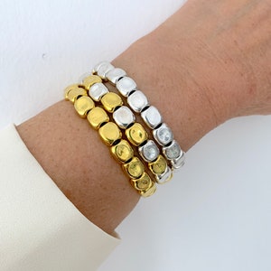 Può includere: Tre braccialetti con perline quadrate in oro e argento. I braccialetti sono indossati su un polso. Il braccialetto dorato è in basso, quello argentato in alto e un braccialetto oro e argento al centro. Le perline misurano circa 1 cm.