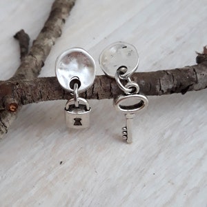 May include: A pair of silver earrings, one with a small padlock charm and the other with a small key charm. The charms are hanging from round silver discs.