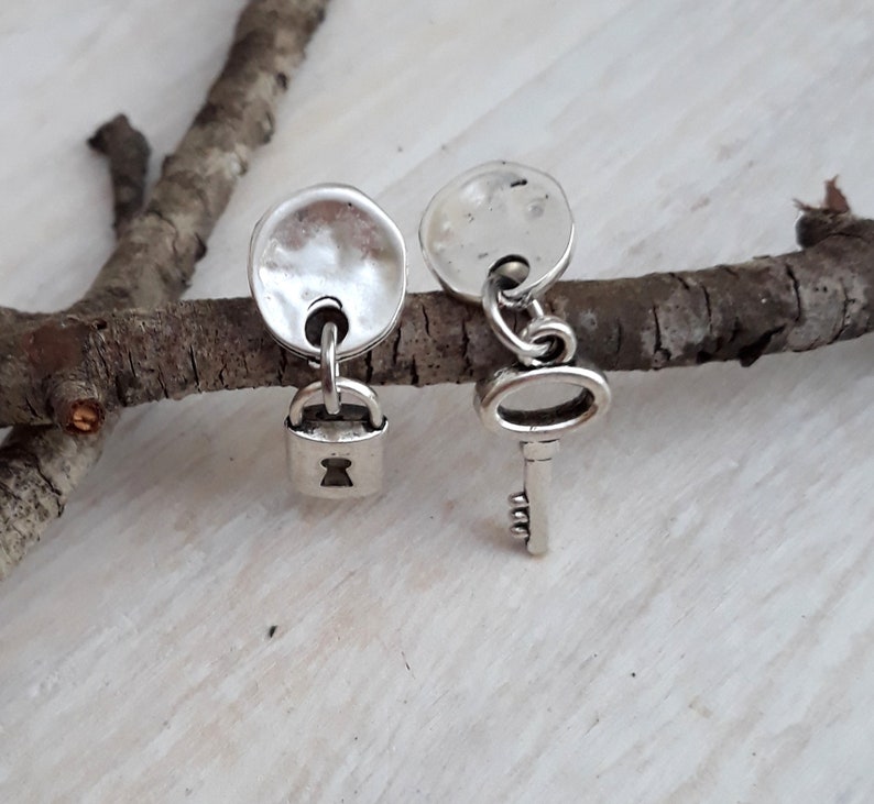 May include: A pair of silver earrings, one with a small padlock charm and the other with a small key charm. The charms are hanging from round silver discs.