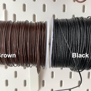 K&ouml;nnte beinhalten: Zwei Spulen Lederkordel, eine braune und eine schwarze, werden nebeneinander gezeigt. Die braune Kordel ist mit "Brown" und die schwarze Kordel mit "Black" beschriftet. Die Kordeln sind eng auf wei&szlig;en Spulen aufgewickelt.