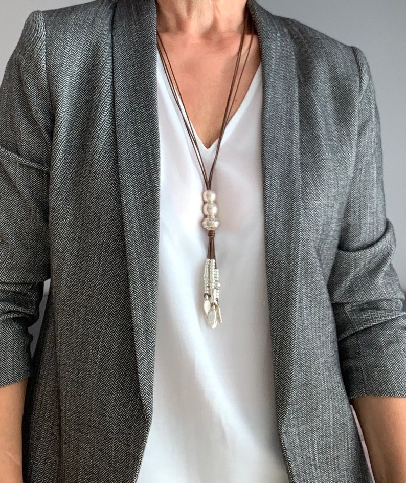K&ouml;nnte beinhalten: Eine lange, braune Lederkette mit silbernen Perlen und einem Quastenanh&auml;nger. Die Kette wird &uuml;ber einem wei&szlig;en Oberteil und einem grauen Fischgr&auml;t-Blazer getragen.