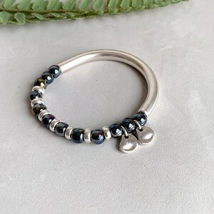 Pode incluir: Um bracelete de prata com contas pretas iridescentes e dois pingentes de prata.