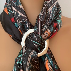 Può includere: Un foulard a motivi neri, verde acqua, arancioni e bianchi, fissato con un anello in metallo color argento liscio. Il tessuto presenta complessi motivi geometrici e floreali su tutta la superficie, tenuti insieme dalla chiusura circolare.