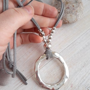 May include: A silver pendant necklace with a large, hammered, circular pendant. The necklace is made with a gray suede cord and silver beads.