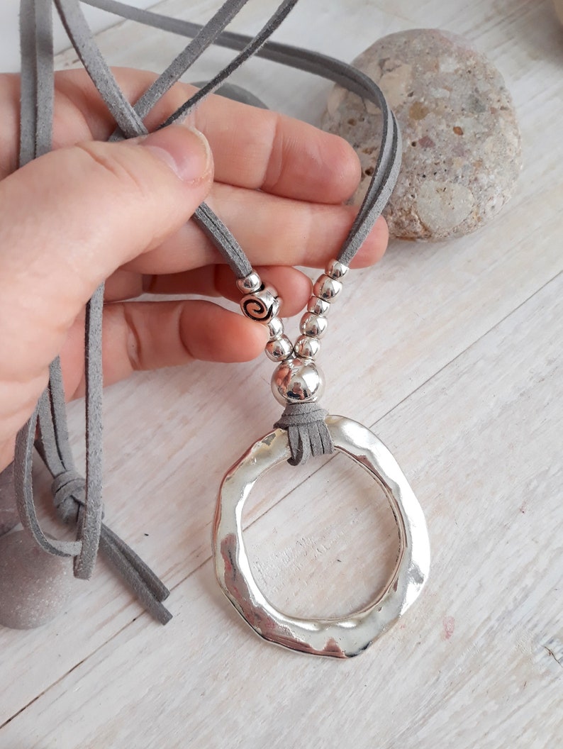 May include: A silver pendant necklace with a large, hammered, circular pendant. The necklace is made with a gray suede cord and silver beads.