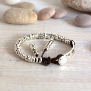 Könnte beinhalten: Ein silbernes Armband mit Perlen, einem braunen Lederband und einer silbernen Platte. Das Armband liegt auf einer weißen Holzoberfläche.