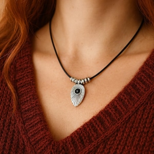 Könnte beinhalten: Halskette mit silberfarbenem Anhänger und schwarzem Band. Der Anhänger hat eine tropfenförmige Form mit schwarzem Zentrum und Sonnenstrahlendesign. Die Halskette wird über einem weinroten Pullover getragen.