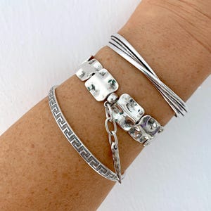 Op de afbeelding: Een verzameling zilveren armbanden. Eén armband heeft een Grieks sleutelontwerp, een andere heeft vierkante, getextureerde schakels en een derde heeft een gedraaid ontwerp. De armbanden worden op een arm getoond.