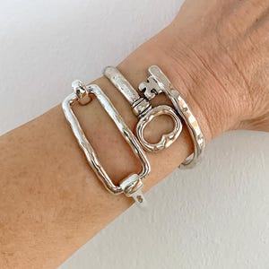 Könnte beinhalten: Ein silbernes Armband mit zwei Anhängern. Ein Anhänger ist ein Rechteck mit einer strukturierten Oberfläche. Der andere Anhänger ist ein Schlüssel mit einem herzförmigen Schlüsselloch.