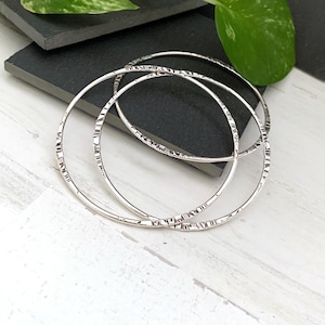 May include: Three silver bangles with a textured, etched design. The bangles are circular and lie on a dark gray surface, with a white wooden surface in the foreground. Green leaves are visible in the background.