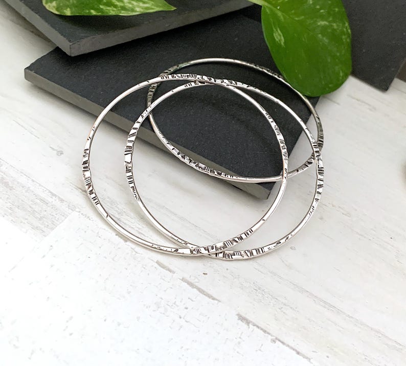 May include: Three silver bangles with a textured, etched design. The bangles are circular and lie on a dark gray surface, with a white wooden surface in the foreground. Green leaves are visible in the background.