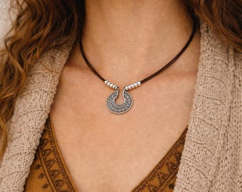 May include: A silver pendant necklace with a brown cord. The pendant is a half-moon shape with intricate etched designs. The necklace is worn over a brown top and a beige cardigan.