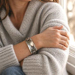 Può includere: Bracciale argentato con centro rettangolare e dettagli strutturati. Il bracciale è indossato su un polso, con un maglione grigio chiaro e jeans blu sullo sfondo. Il bracciale sembra essere in metallo.