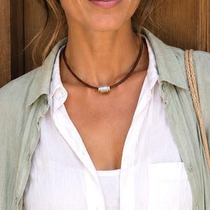May include: A brown leather cord necklace featuring a single cylindrical silver-toned metal bead at the center. The necklace rests against the neck, showcasing a smooth, dark brown leather band and a polished, ribbed metallic accent piece.