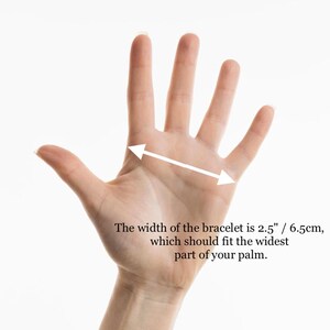 May include: A hand with a white arrow indicating the width of a bracelet. The text states the width is 2.5 inches / 6.5cm, which should fit the widest part of your palm.