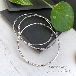 May include: Three silver-plated bangles with textured detailing. The bangles are circular and rest on a dark gray surface. The text "Silver plated (not solid silver)" is visible in the image.