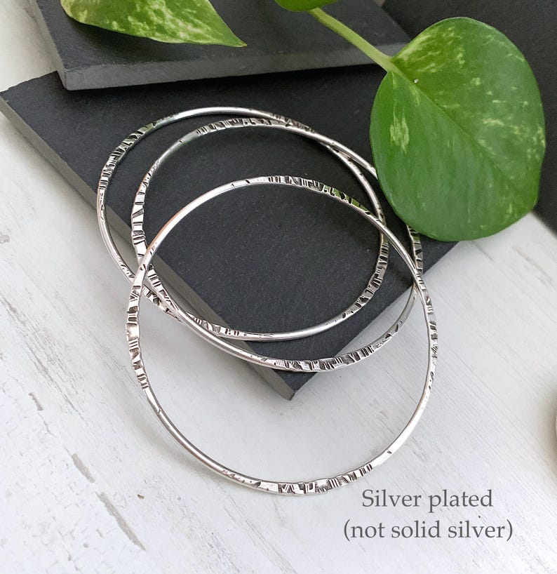 May include: Three silver-plated bangles with textured detailing. The bangles are circular and rest on a dark gray surface. The text "Silver plated (not solid silver)" is visible in the image.