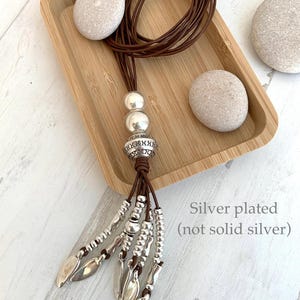 K&ouml;nnte beinhalten: Eine lange braune Lederkette mit versilberten Perlen und blattf&ouml;rmigen Anh&auml;ngern. Die Kette hat einen dekorativen silbernen Perlenabschnitt und wird auf einem Holztablett mit glatten Steinen pr&auml;sentiert. Text: Versilbert (nicht massiv Silber).