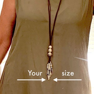 K&ouml;nnte beinhalten: Eine lange braune Halskette mit silberfarbenen Perlen und einem dekorativen Anh&auml;nger h&auml;ngt &uuml;ber einem &auml;rmellosen olivgr&uuml;nen Kleid. Der Text "Your size" ist mit Pfeilen auf dem Bild geschrieben, die auf die Halskette zeigen.